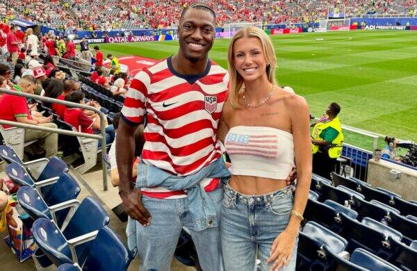 Smiling couple poses together in a stadium: man in a red-and-white USA jersey with denim jacket tied around the waist, woman in a white crop top with a US flag, both wearing jeans; green field behind them.