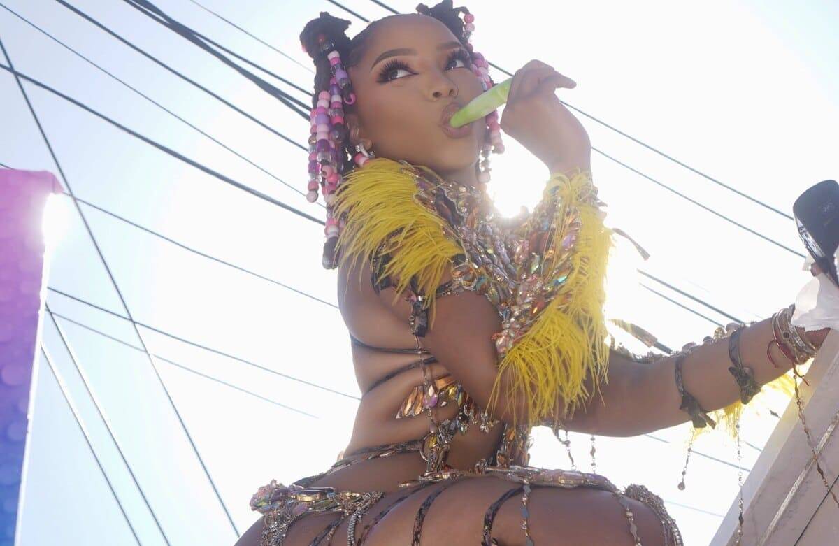 Performer in a bright festival outfit with yellow feathers, licking a green popsicle on stage.