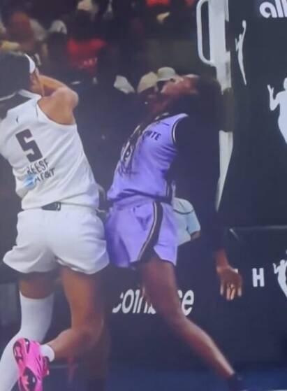 Basketball players in white (number 5) and purple uniforms collide at the sideline during a game near advertising boards.