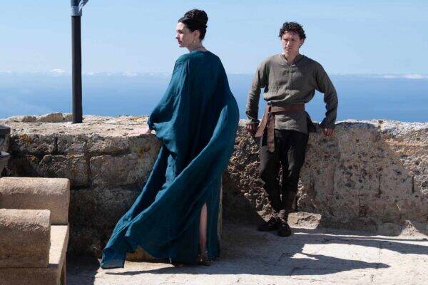 Two people stand at a stone railing overlooking the sea: a woman in a flowing teal cloak facing away, and a man in a gray tunic with a belt.