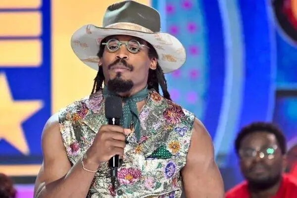 Male performer in a floral vest and wide-brim hat speaks into a microphone on a colorful stage set.