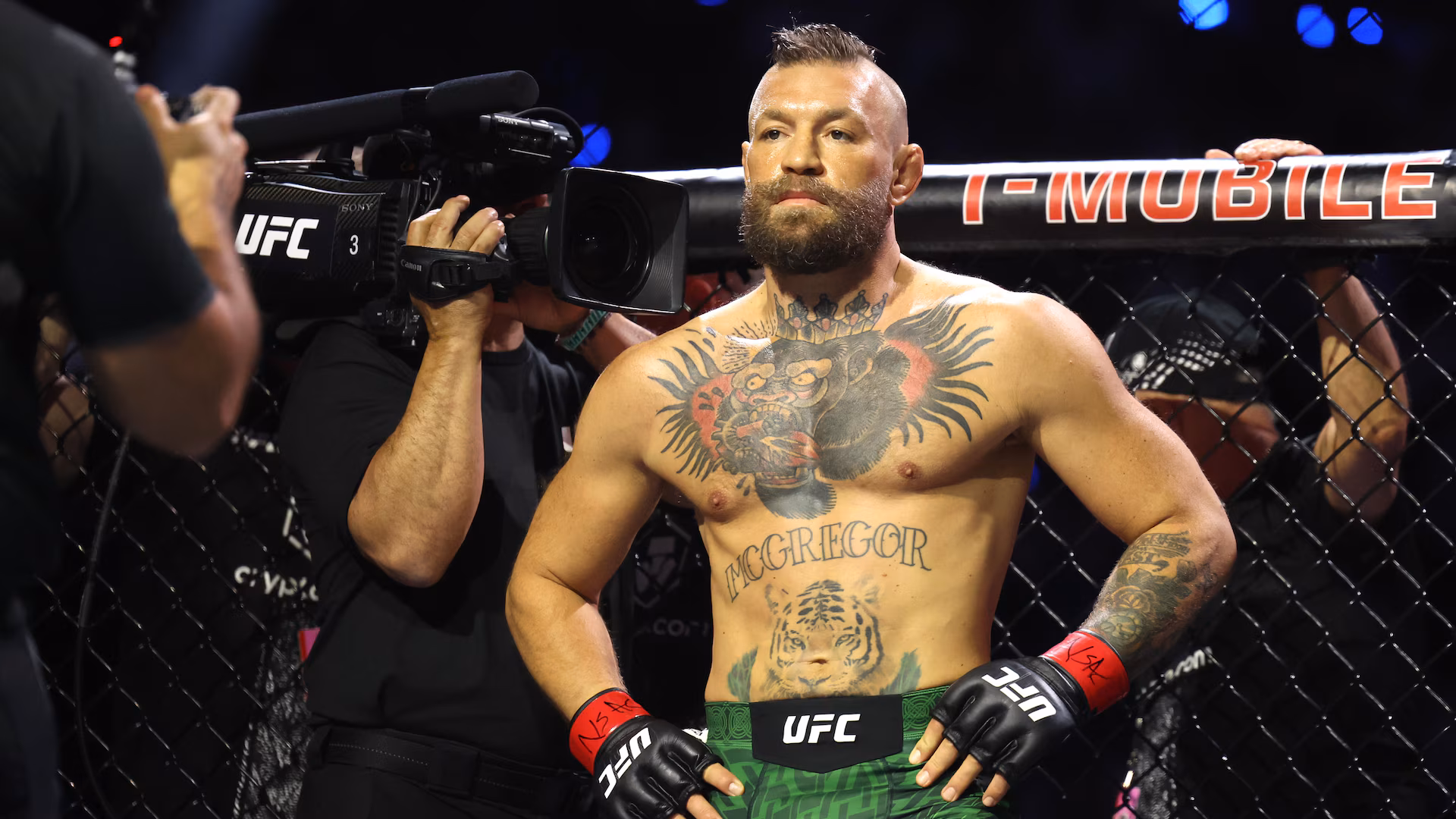 Tattooed UFC fighter in green shorts and red gloves stands in the cage while a cameraman films him from the left.