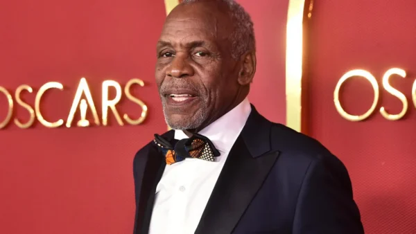 Older man in a black tuxedo and colorful bow tie on a red carpet, with OSCARS signage in the background