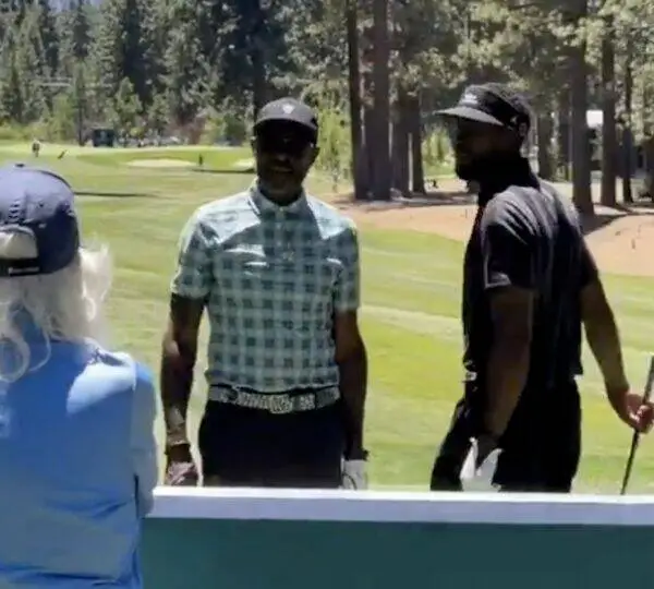 Two men stand on a sunny golf course, one in a checkered shirt and cap, the other in a dark shirt and visor, with trees in the background.