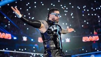 WWE wrestler in a black shiny suit with blue sunglasses raises arms in the ring, lights sparkling around him.