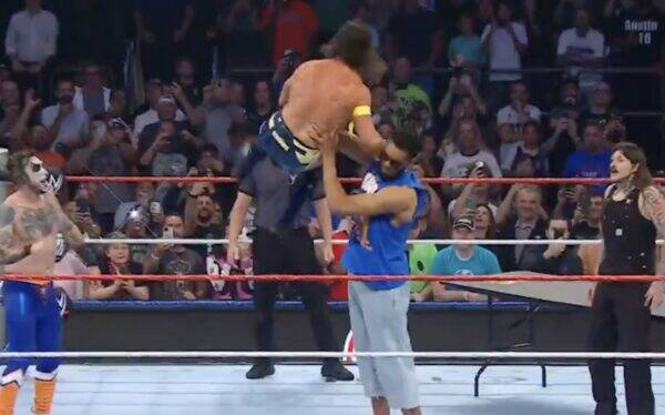 Wrestling match in a crowded arena; masked wrestler in blue pants stands left while a muscular opponent is lifted overhead by a referee or teammate in the center of the ring as spectators film.
