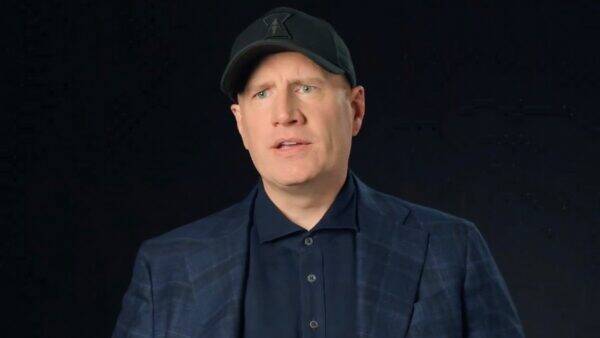 Man in a navy plaid blazer and dark shirt speaks against a solid dark background, wearing a black cap.