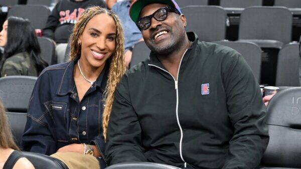 Smiling couple seated in stadium seats; woman with braided hair in a denim jacket, man in a black zip jacket and cap.
