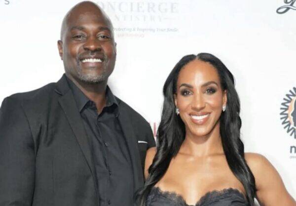 Smiling man in a black suit beside a smiling woman in a black lace dress at a formal event on a white backdrop