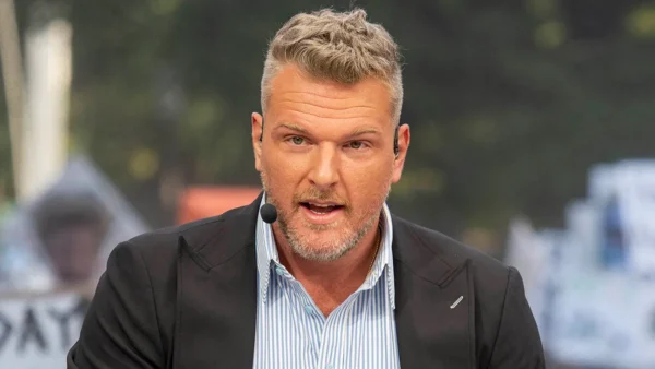 Adult male host wearing a headset microphone, speaking to the camera outdoors in a black blazer and striped shirt.