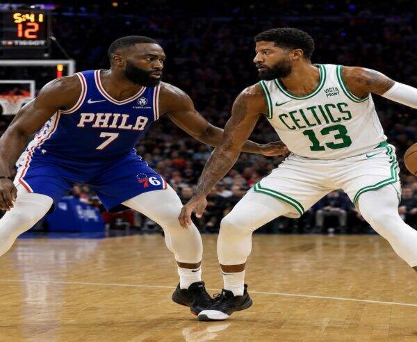 Two basketball players, one in a blue Phillies jersey (number 7) and the other in a white Celtics jersey (number 13), guard each other on a hardwood court during a game, with a scoreboard visible in the background.