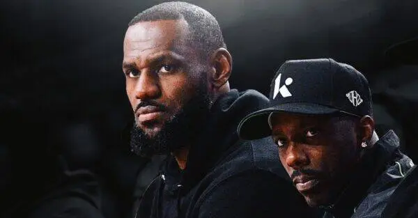 Two men in dark clothing pose closely against a dark background, one with a beard and the other wearing a cap with a white logo on the side.