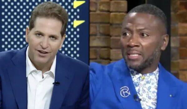 Split-screen of two men in a TV studio: left host in a blue suit speaks, right guest in a bright blue blazer gestures amid a brick background