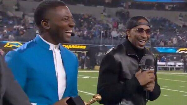 Two men on a football field during a broadcast, one in a bright blue suit and the other in a black jacket with a cap, both smiling.