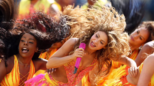 Front-center performer singing into a pink microphone while dancing in a vibrant orange-yellow festival scene with other dancers around her.
