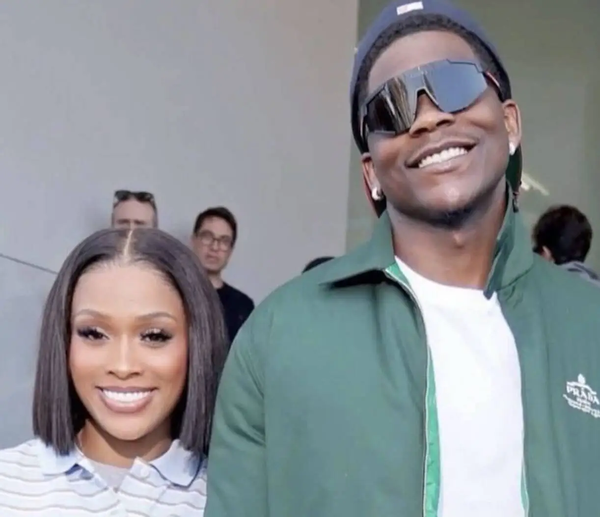 Smiling woman with a straight bob haircut posing beside a man in a green jacket and oversized sunglasses indoors, with people in the background.
