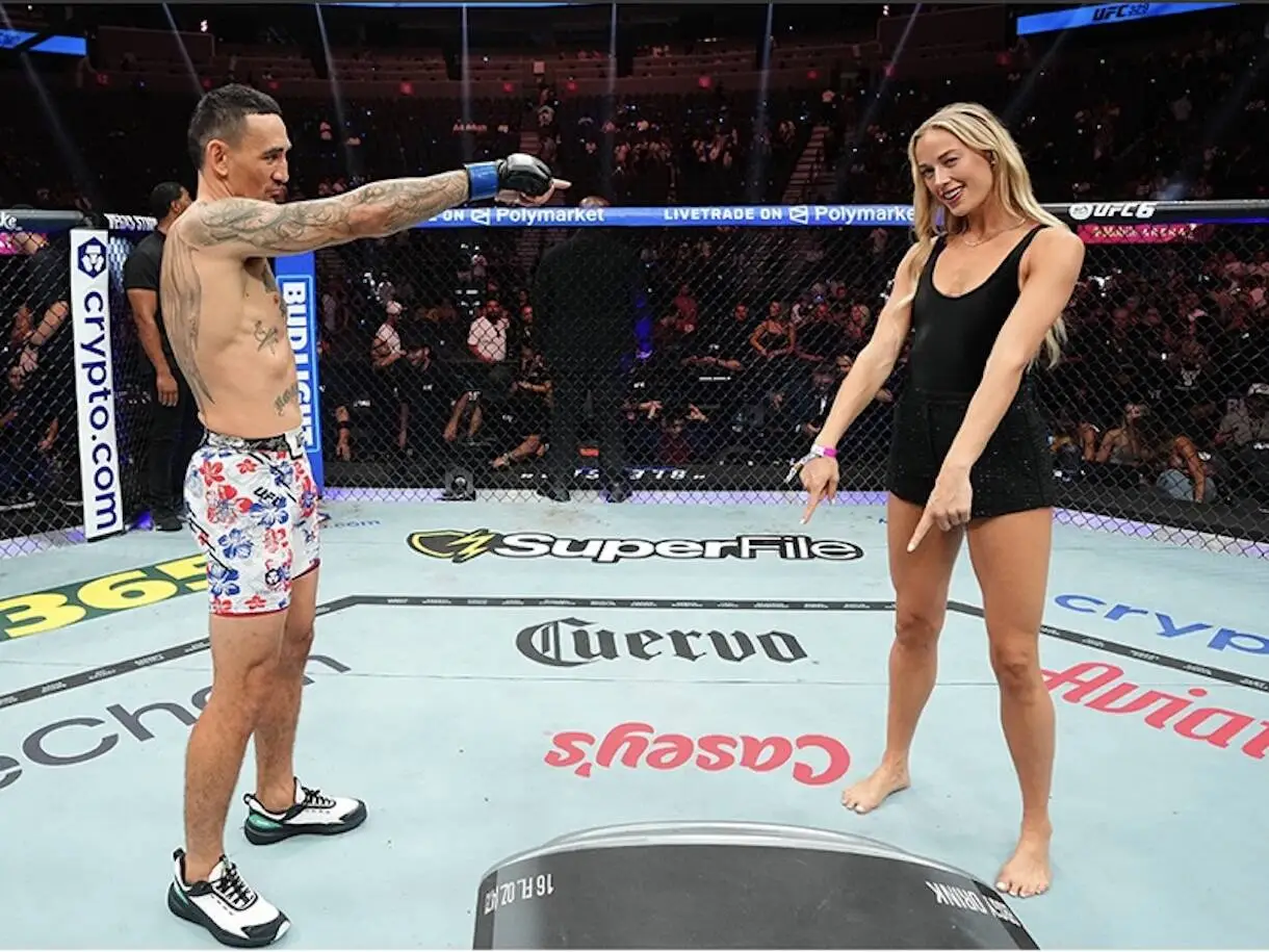 Two people stand in a UFC octagon facing each other; man points toward a woman in a black dress as spectators watch.