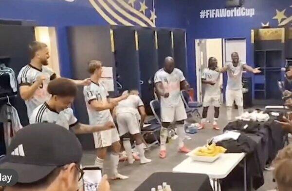 Soccer players in white uniforms celebrate in a locker room, several standing with arms raised and clapping near blue walls with #FIFA World Cup.