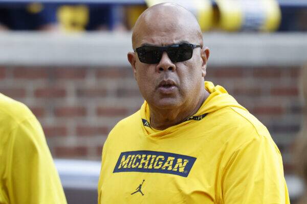 Man in a bright yellow Michigan shirt and dark sunglasses, speaking or mid-sentence outdoors.