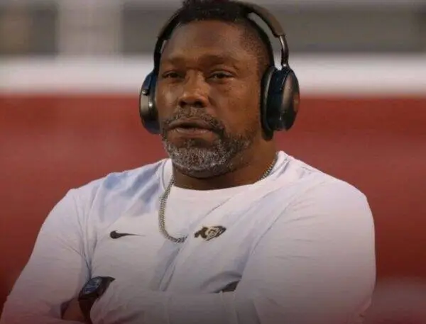 Man wearing large over-ear headphones and a white Nike shirt, arms crossed, in a stadium setting.