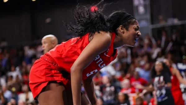 Female basketball player in a red uniform leans forward with a fierce expression during a game, crowd in the stands blurred behind her.