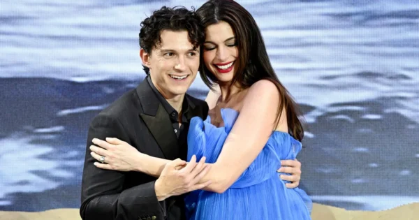 A smiling couple embraces on stage, the man in a black suit and the woman in a blue dress, backdrop of ocean scenery behind them.