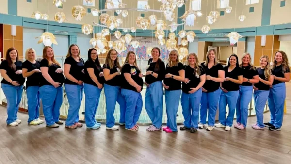 Group of pregnant women posing in a hospital lobby, wearing black tops and blue pants with hands on bellies.