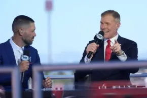 Two men in navy suits with FOX Sports mics conversing on a broadcast stage outdoors.