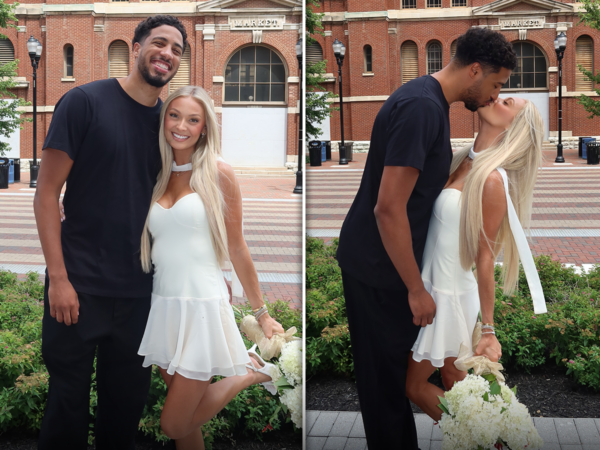 Couple posing outside a red brick market building; man in black, woman in a white dress holding white flowers.