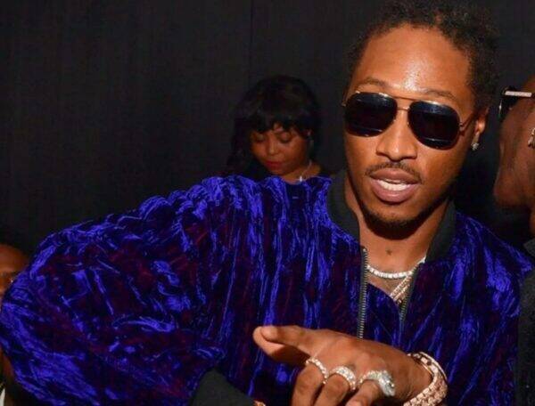 Man wearing a purple velvet jacket and sunglasses, displaying diamond rings while gesturing at a party.