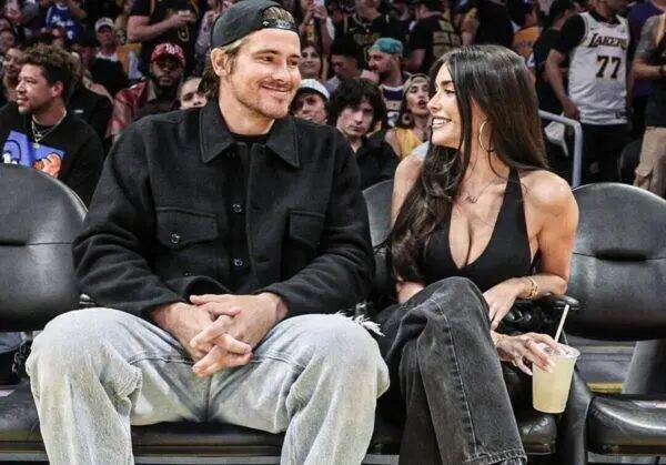 Couple sitting courtside at a sports event, smiling at each other; man in a black jacket and cap, woman in a black top holding a drink.