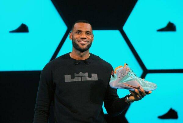 Man on a stage holding a teal sneaker with orange accents, against a bright blue geometric backdrop.