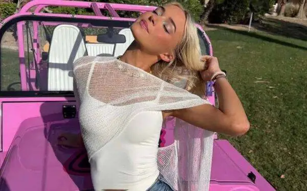 Woman leaning back with eyes closed, posing atop a bright pink convertible in a park. She wears a white top and sheer shawl, styling a relaxed sunny moment.