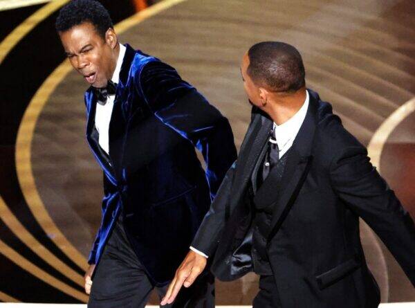 Two men in formal tuxedos dance on a stage with a circular gold backdrop behind them.