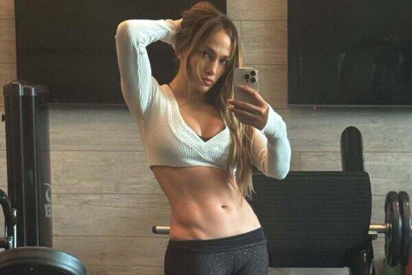 Fitness model in a gym taking a mirror selfie with a smartphone, wearing a white cropped sweater and black leggings, displaying toned abs.