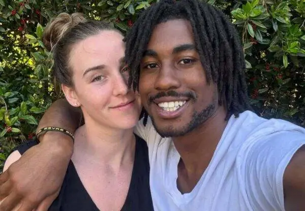 Two friends or a couple pose for a selfie outdoors, smiling with their arms around each other in front of green bushes.