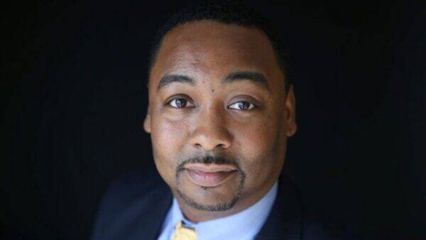 Close-up headshot of a man in a navy suit and yellow tie, looking at the camera against a dark background.