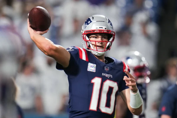Patriots quarterback in navy jersey (#10) throwing a pass during a game, stadium crowd blurred in the background.