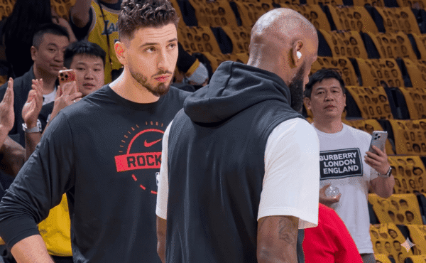 Two basketball players converse on a court; one faces the camera in a black shirt with a red logo, the other has his back turned in a white T-shirt with a vest.