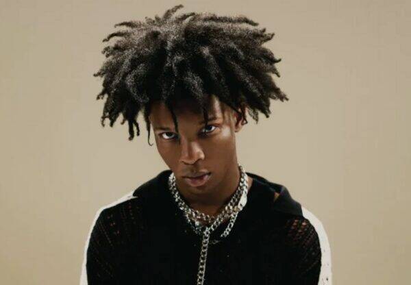 Portrait of a young man with a large afro, wearing a black knit shirt and chunky chains, gazing intently at the camera.