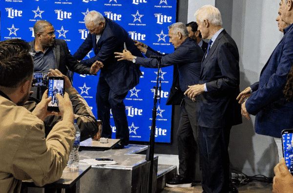 Men in suits on a stage with a blue sponsor backdrop; one man is being helped up as others reach out, while photographers record the moment.