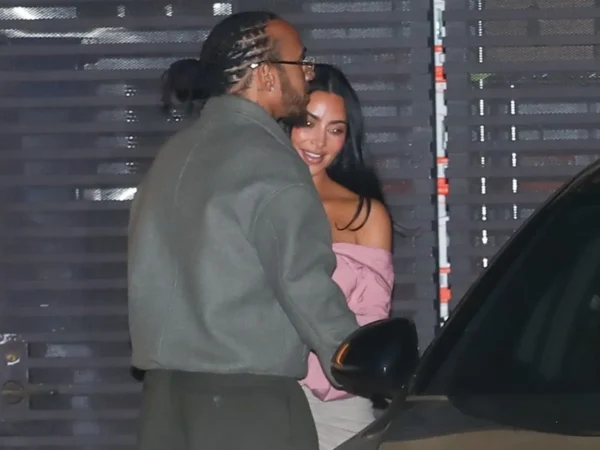 Man with braided hair and beard in a gray coat standing beside a smiling woman wearing a pink off-the-shoulder top near a car at night.