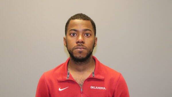 Mugshot-style photo of a man in a red Oklahoma jacket against a plain gray background.