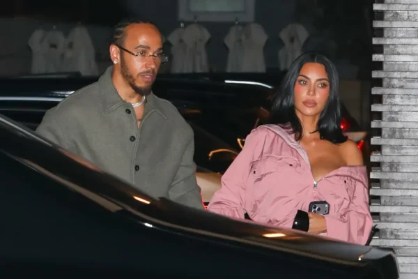 A man and a woman exit a dark car at night; the woman wears a pink off‑shoulder jacket and holds a phone, the man wears a gray coat and glasses.