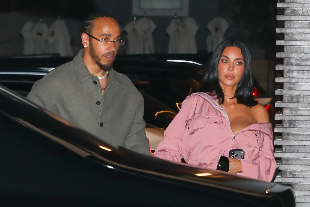 A man and a woman exit a dark car at night; the woman wears a pink off‑shoulder jacket and holds a phone, the man wears a gray coat and glasses.