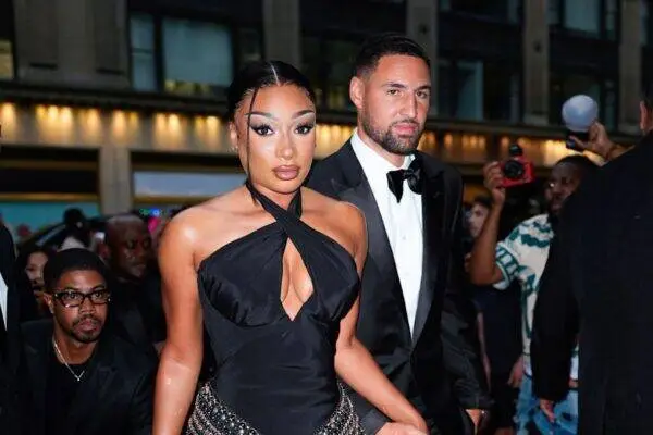 Glamorous couple in formal wear walking a red carpet; woman in a black halter gown with a front cutout and jeweled belt, man in a black tuxedo behind her.
