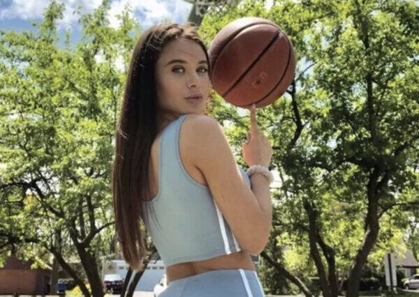 Woman in a light blue athletic outfit outdoors, spinning a basketball on her finger with trees in the background.