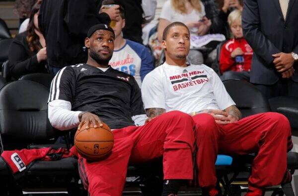 Two Miami Heat players sit courtside in team warmups; one holds a basketball and looks upward while the other gazes ahead, with fans in the background.