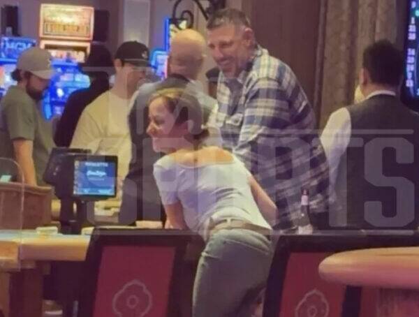 Woman leaning on a casino bar, smiling, with a tall man in a plaid shirt beside her and others in the background.