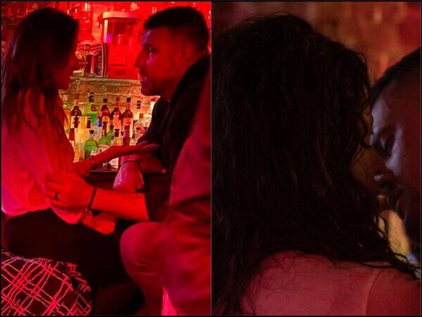 Two people sit at a red-lit bar, facing each other and holding hands in a close, intimate conversation at nightlife venue.